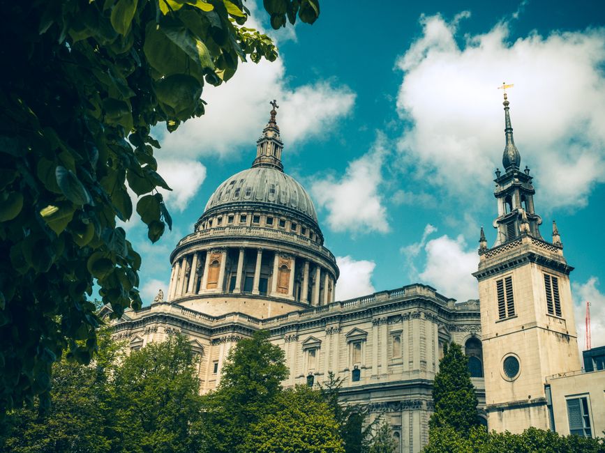 A historical landmark with a large classic dome topped by a small spire, featuring ornate architectural details, flanked by two towers with peaked roofs and clock windows. The building is surrounded b