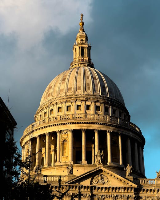 A close-up view of St. Paul's Cathedral's iconic dome with intricate architectural details including columns and sculptures at its base, set against a partly cloudy sky with sunlight illuminating the 