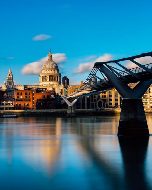 A close-up view of St. Paul's Cathedral's iconic dome with intricate architectural details including columns and sculptures at its base, set against a partly cloudy sky with sunlight illuminating the 
